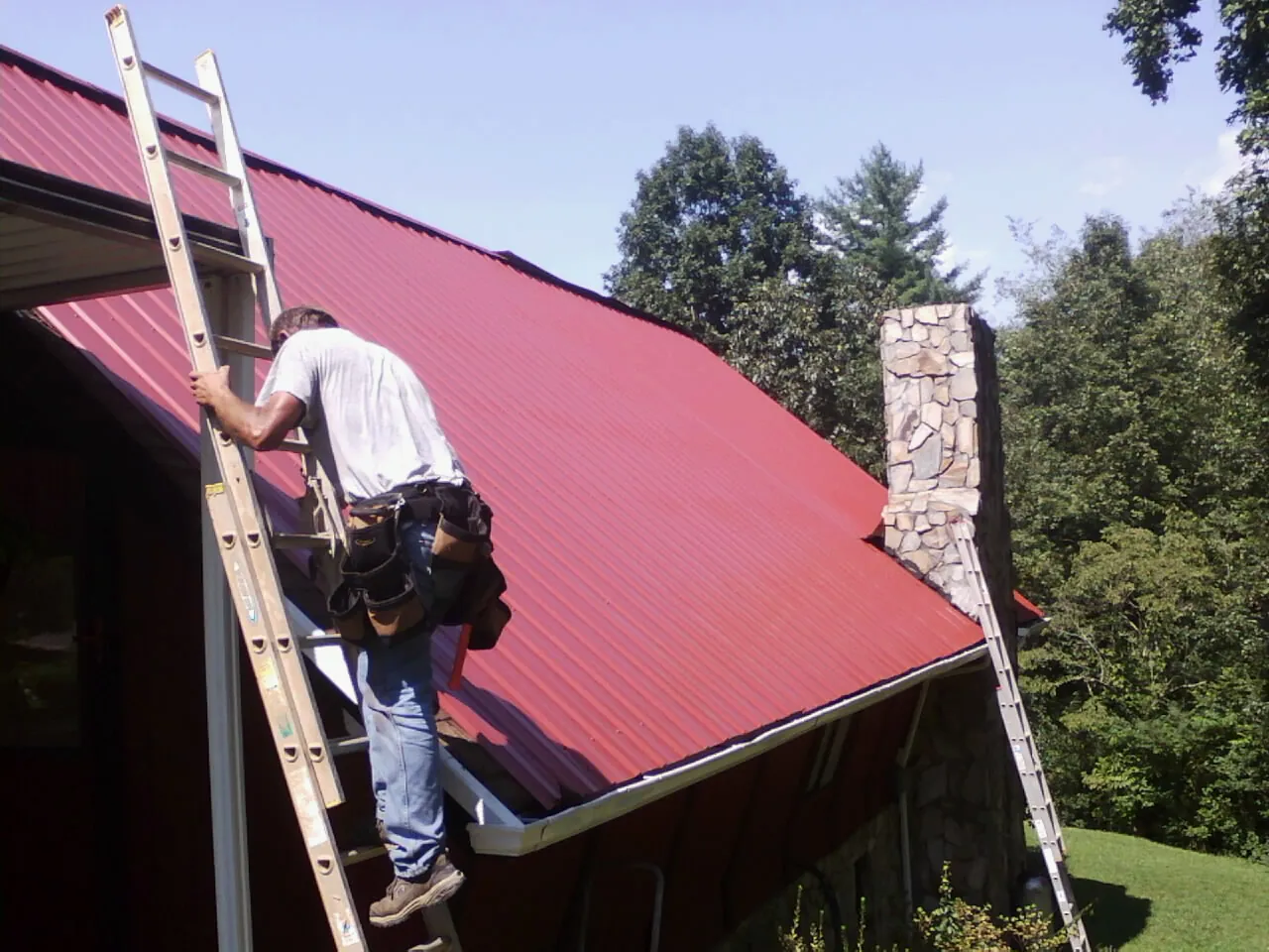 Roofer on a ladder beside a red metal roof and stone chimney — Soffit & Fascia Repair by Roofing Brothers in Fiatt